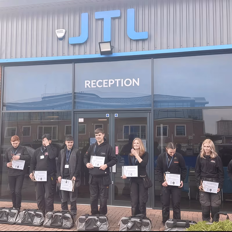 Seven people standing in a row outside a building with JTL signage and a reception door, holding certificates and black bags on the ground in front of them.