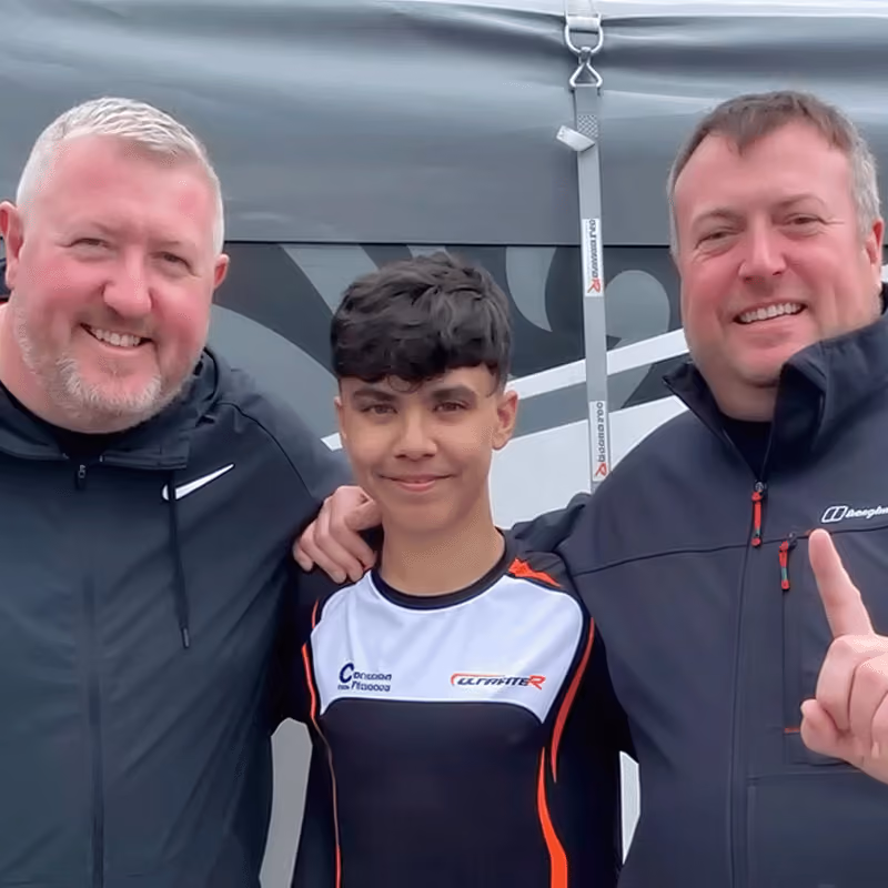Three smiling people stand close together in front of a tent, with the person on the right holding up one finger and the younger person in the middle wearing a racing uniform shirt.