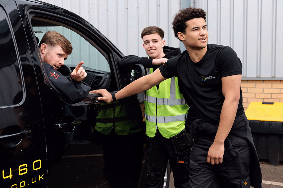 Three young team mebers in work uniforms near a company van; one man leans out of the driver side window pointing, another in a neon safety vest stands by the vehicle, and a third man leans on the vehicle looking ahead.
