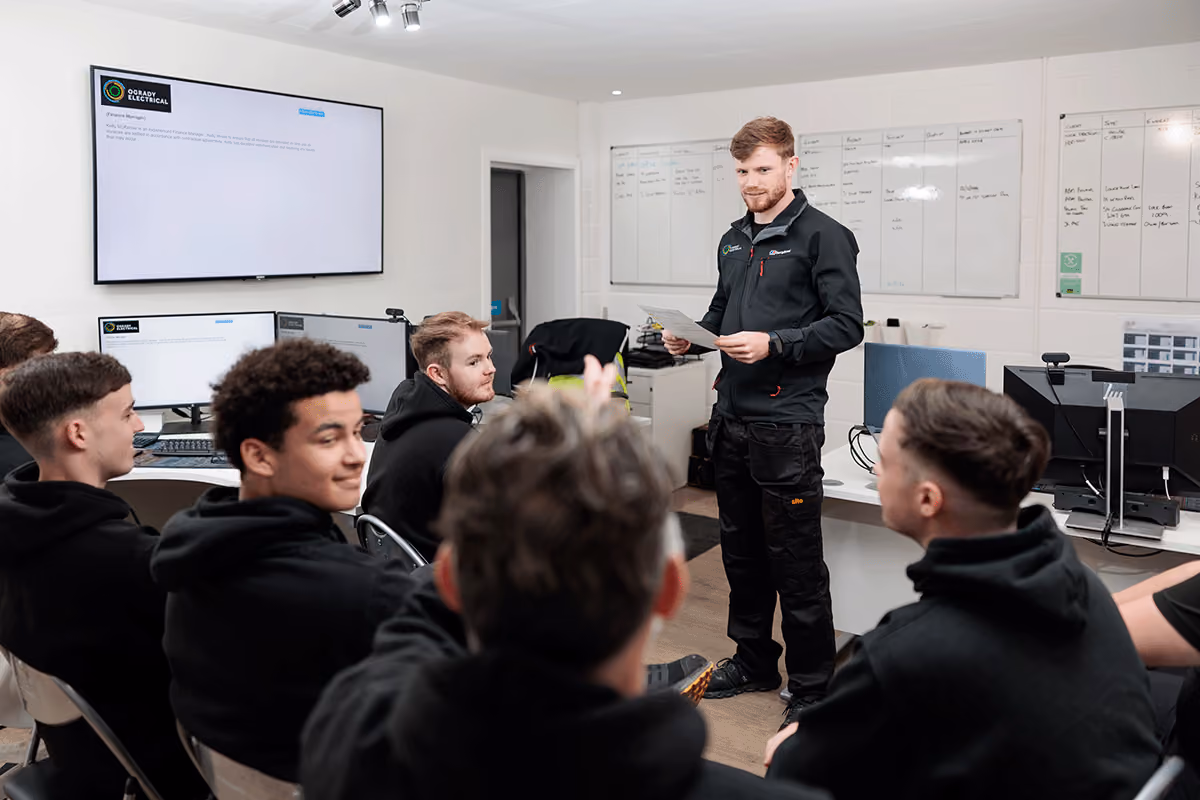 An O'grady team lead standing and addressing a group of six young men seated and attentively listening in a modern office with multiple screens and whiteboards during company briefing session.