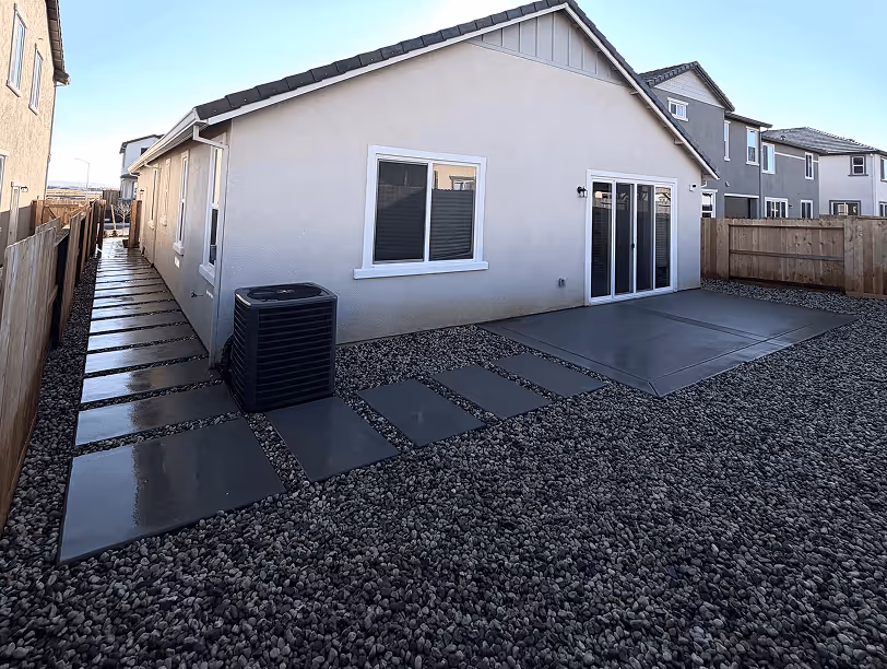 Backyard of a house featuring a gray pebble ground covering, rectangular concrete stepping stones leading to a side path, and a concrete patio adjacent to the house's sliding glass doors.