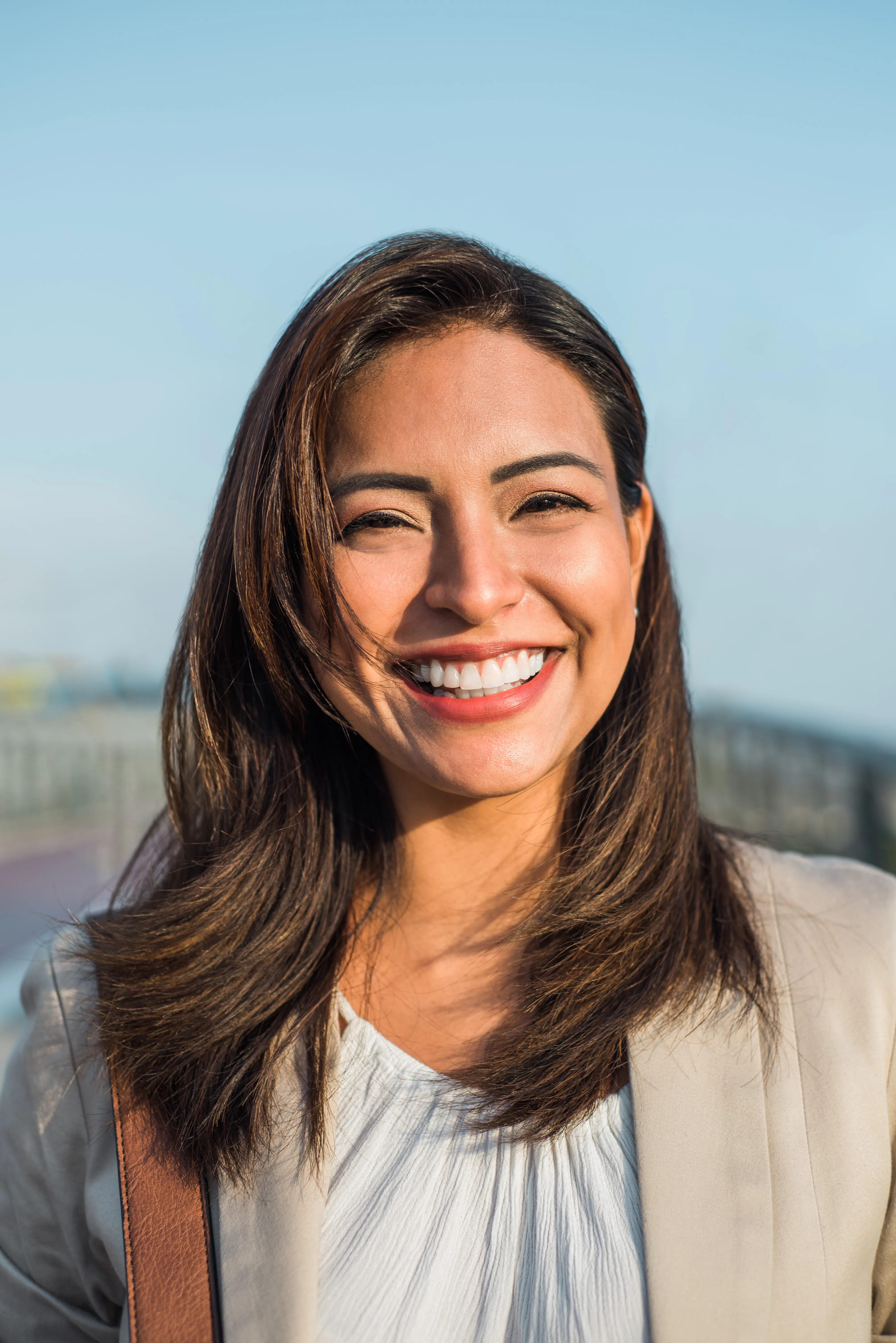 Young woman with long dark hair wearing white top, smiling outdoors with sky visible behind her