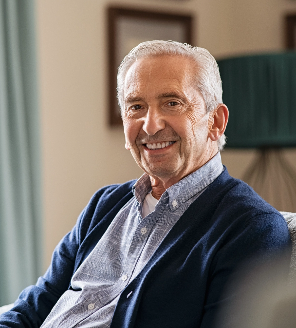 Older man with white hair and stubble wearing gray button-up shirt, smiling indoors with neutral interior background