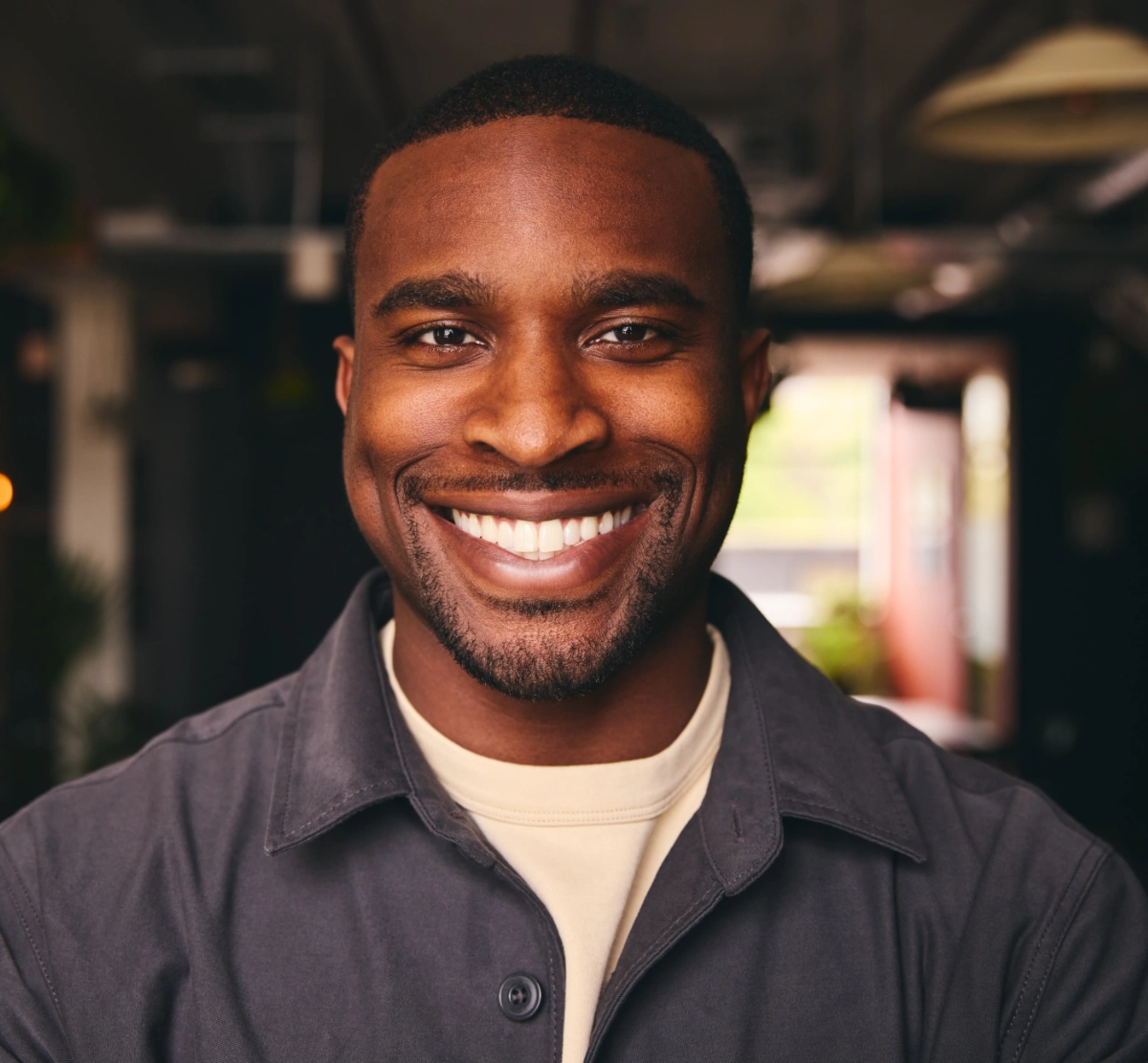 Man with deep skin tone in casual shirt, indoor headshot with warm ambient lighting and blurred warm-toned background