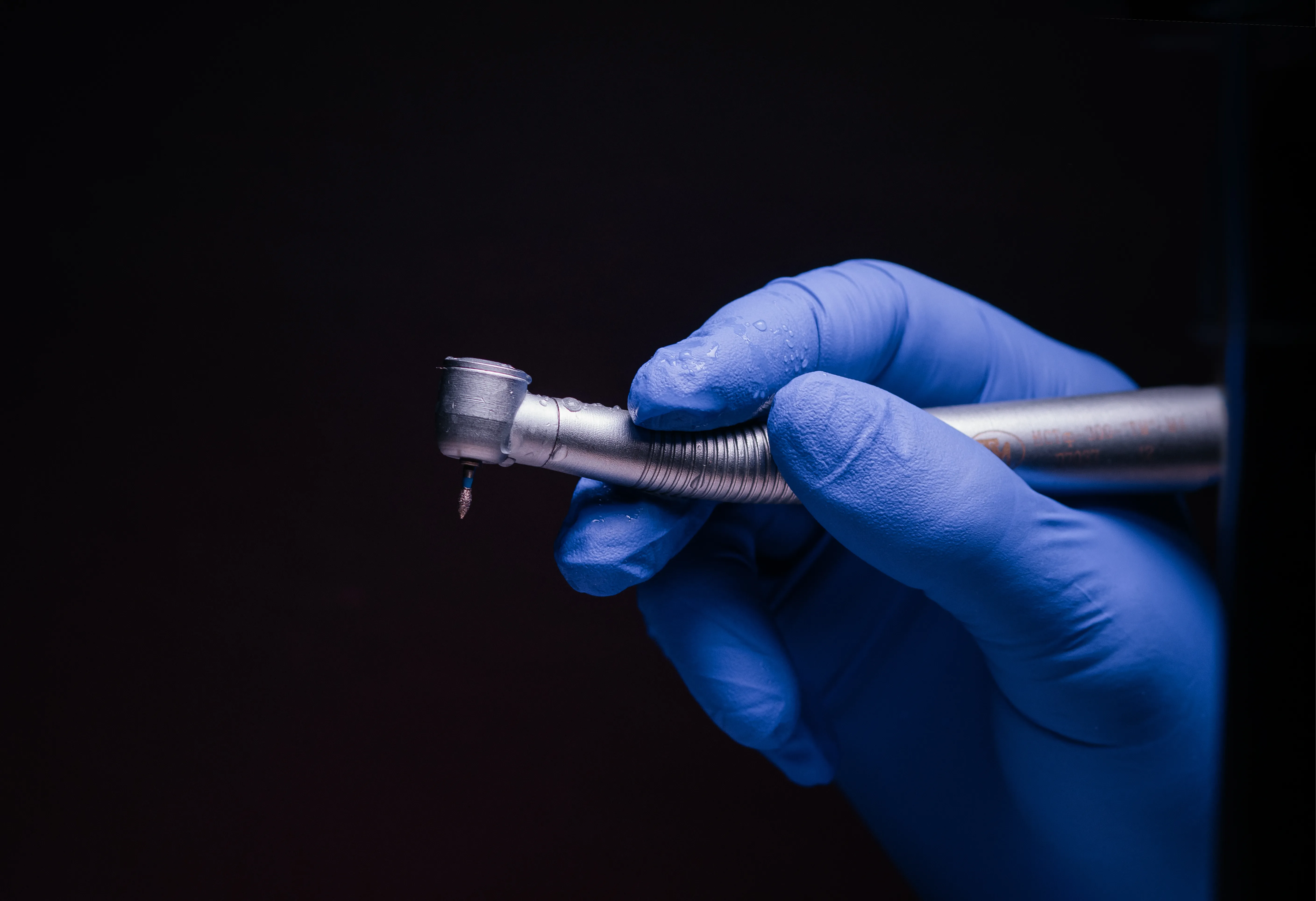 Dental handpiece/drill held in gloved hands against dark blue background with text overlay