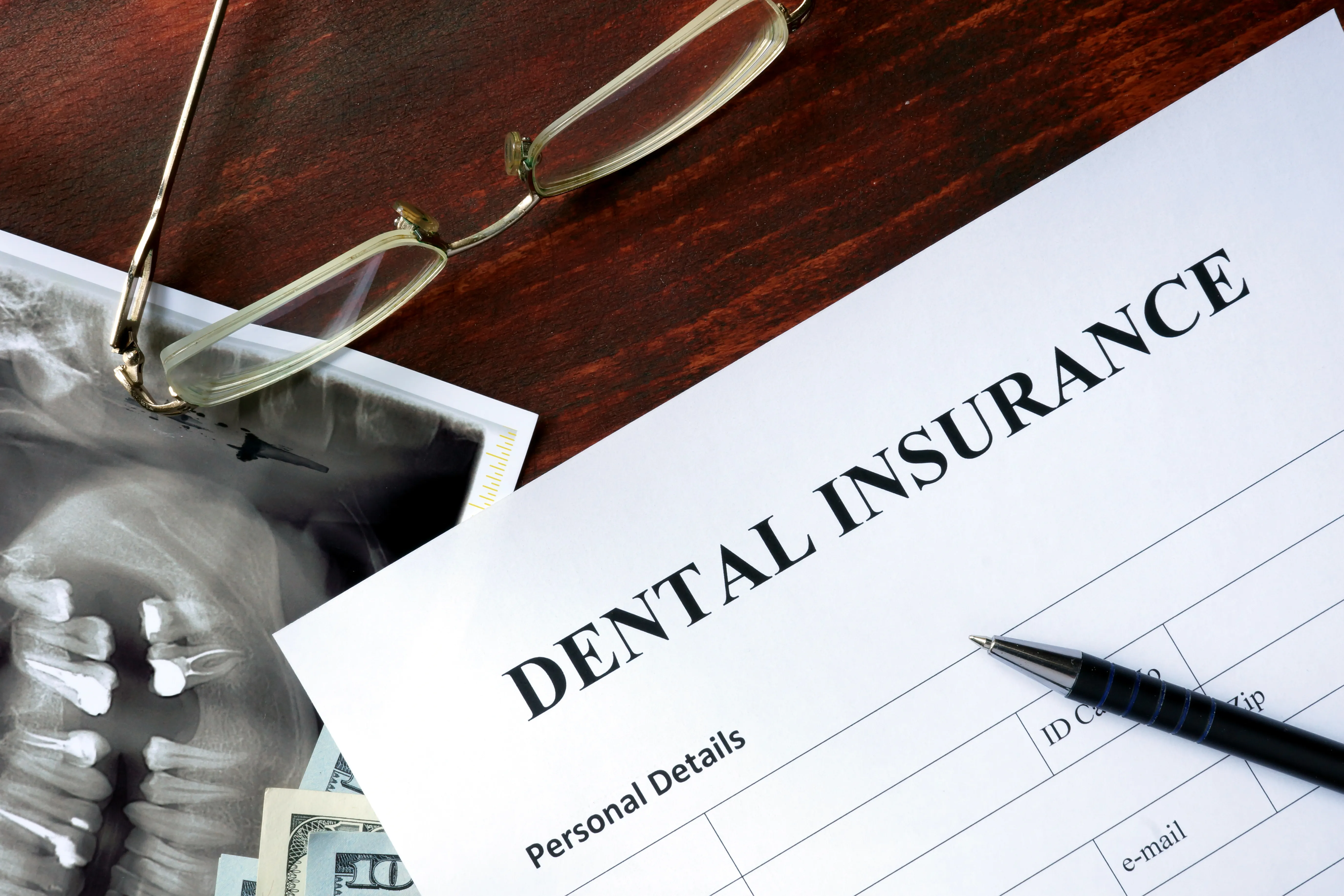 Dental X-ray with glasses and a dental insurance form displaying "DENTAL INSURANCE" text and "Personal Details" section, with pen resting on document
