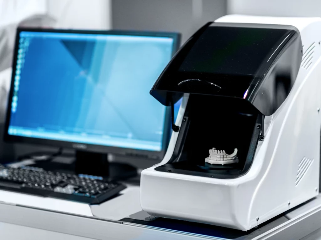 CAD/CAM dental milling machine with white and black components next to computer monitor displaying blue design software