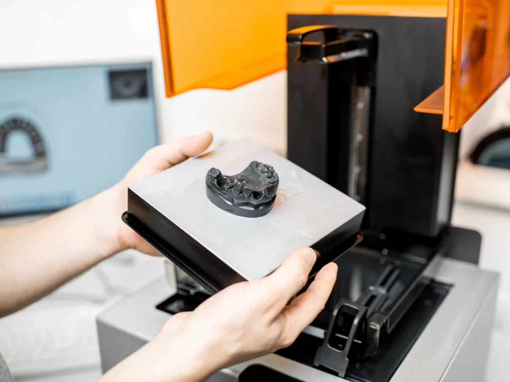 Hands holding white platform with 3D-printed surgical guide or model, with orange and black 3D printer visible in background