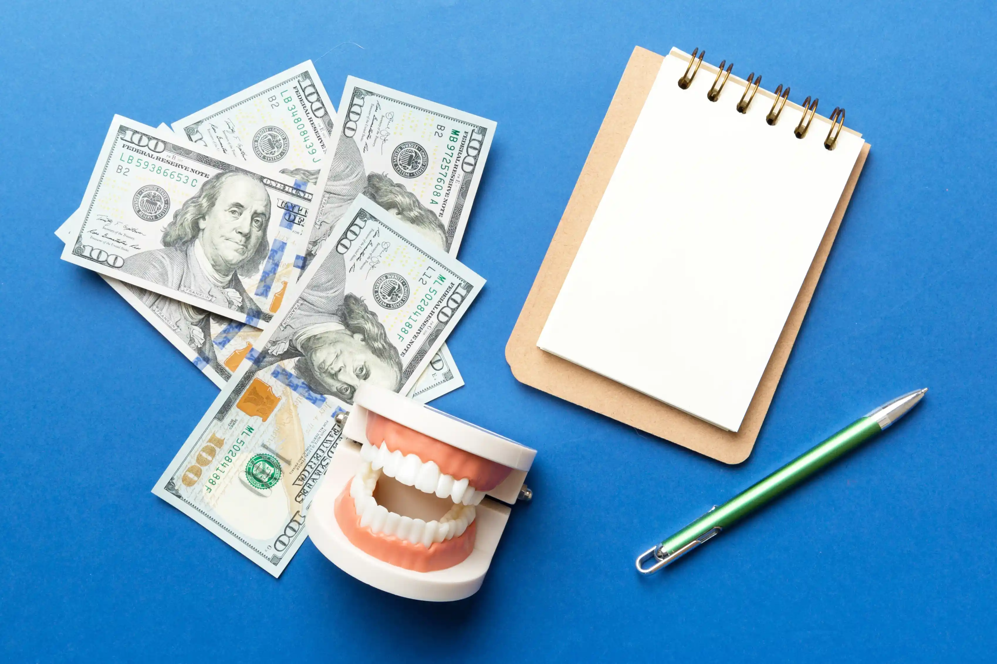 US dollar bills, blank spiral-bound notebook, and dental model showing teeth on blue background with pen