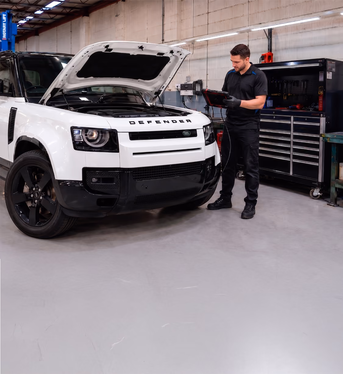 Mechanic using diagnostic tablet on red Land Rover Defender with hood open in a workshop.