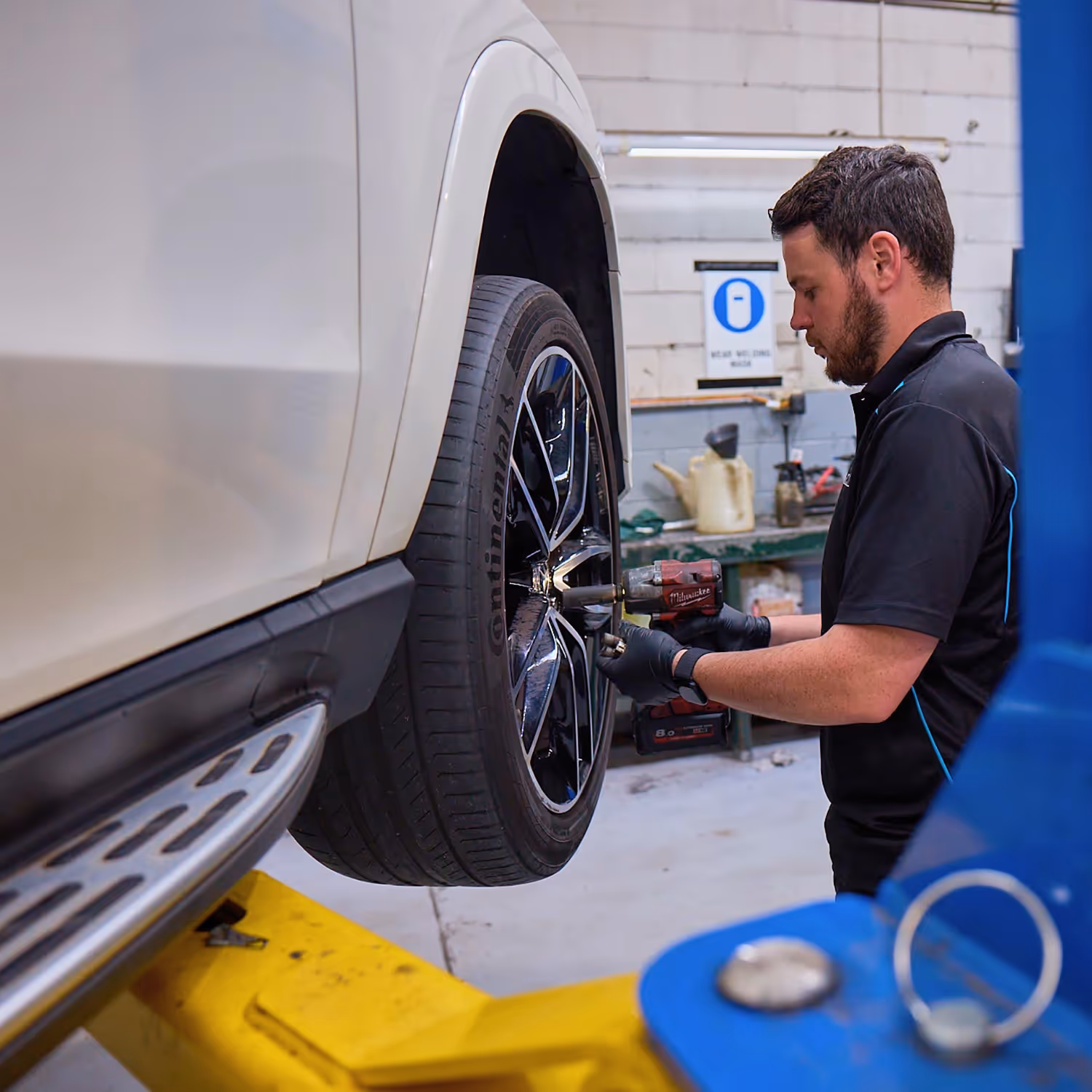 Mechanic using power tool to tighten lug nuts on a black alloy wheel of a white SUV in a garage.