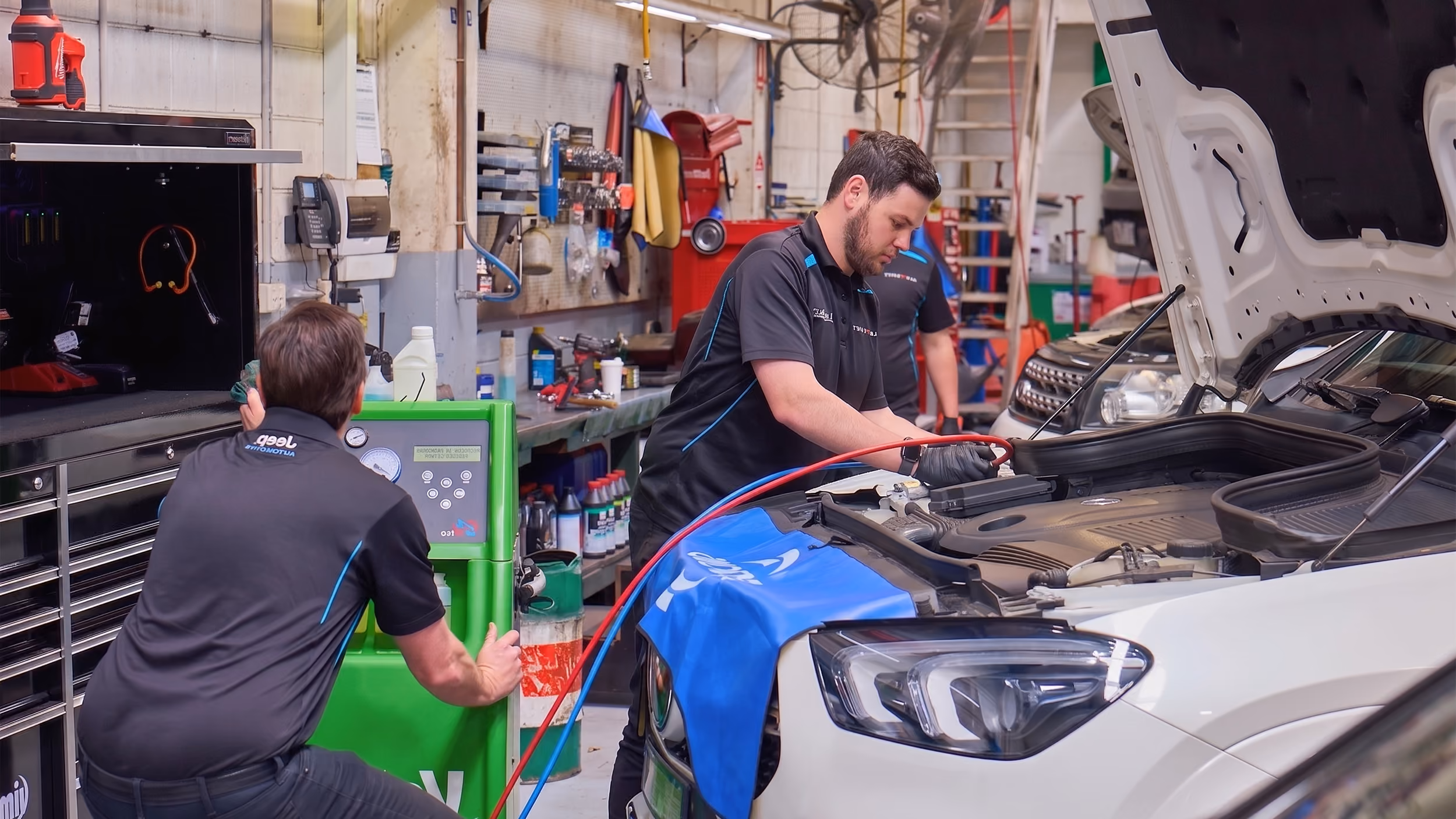 Two mechanics working on a car engine in a busy auto repair shop with diagnostic equipment.