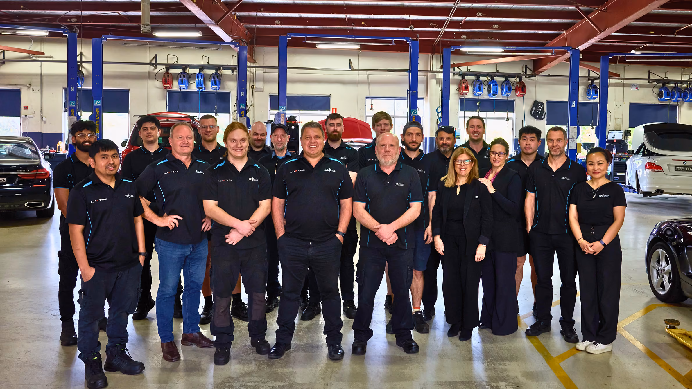 Group of mechanics and staff standing in an auto repair shop with cars and lifts in the background.