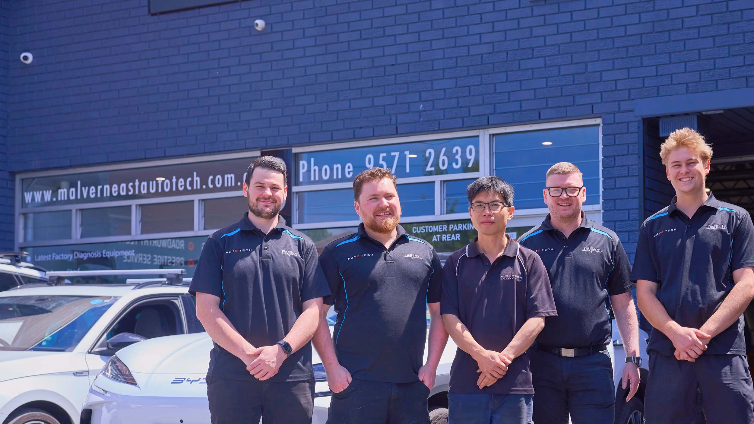 Five men in black Auto Tech shirts stand smiling in front of a white car and building with signage.