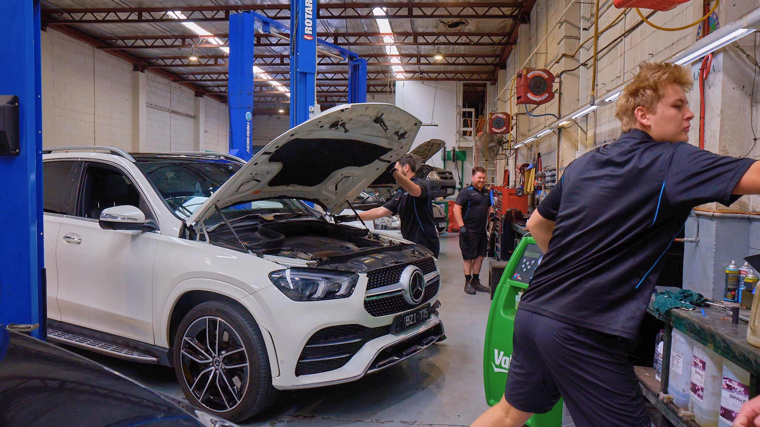 Mechanics working on a white Mercedes SUV in a busy Melbourne workshop during a car inspection.