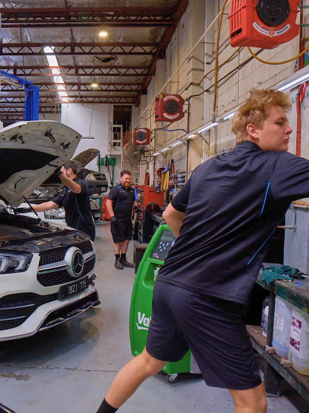 Mechanics working on a white Mercedes SUV in a busy Melbourne workshop during a car inspection.