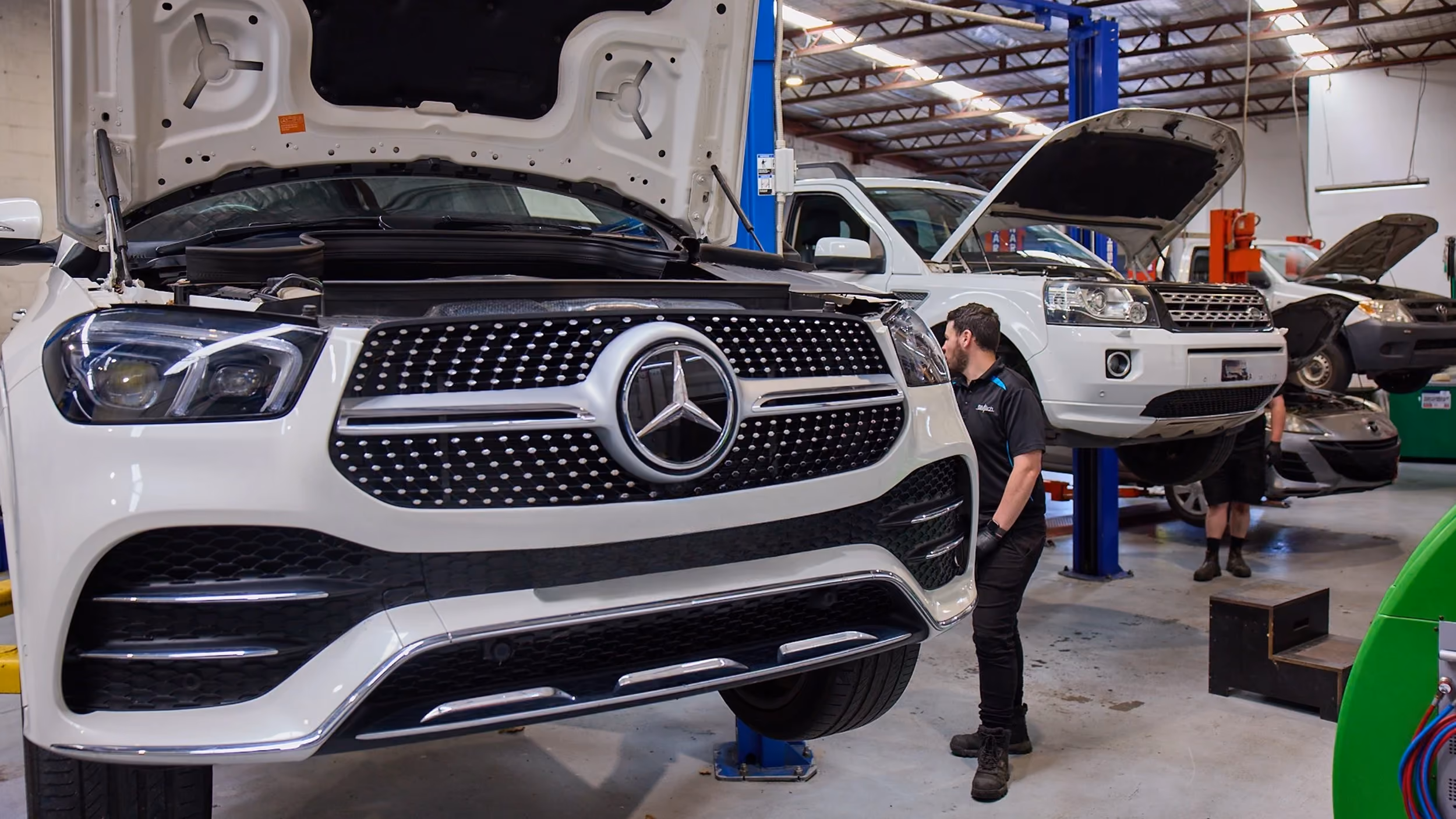 Mechanic inspecting a white Mercedes SUV on a hoist inside a workshop.]