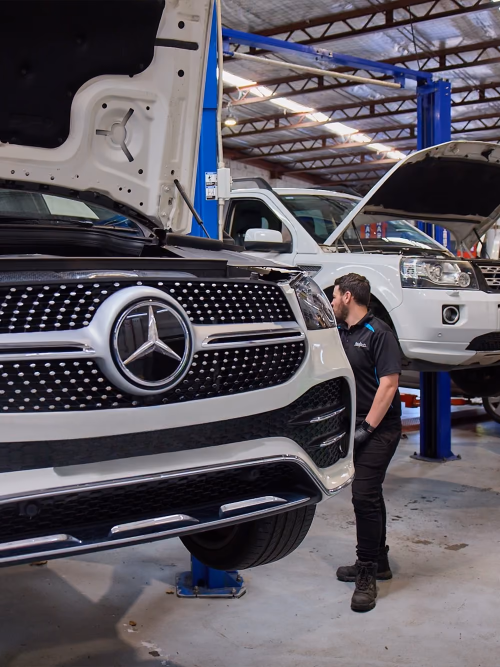 Mechanic inspecting a white Mercedes SUV on a hoist inside a workshop.]
