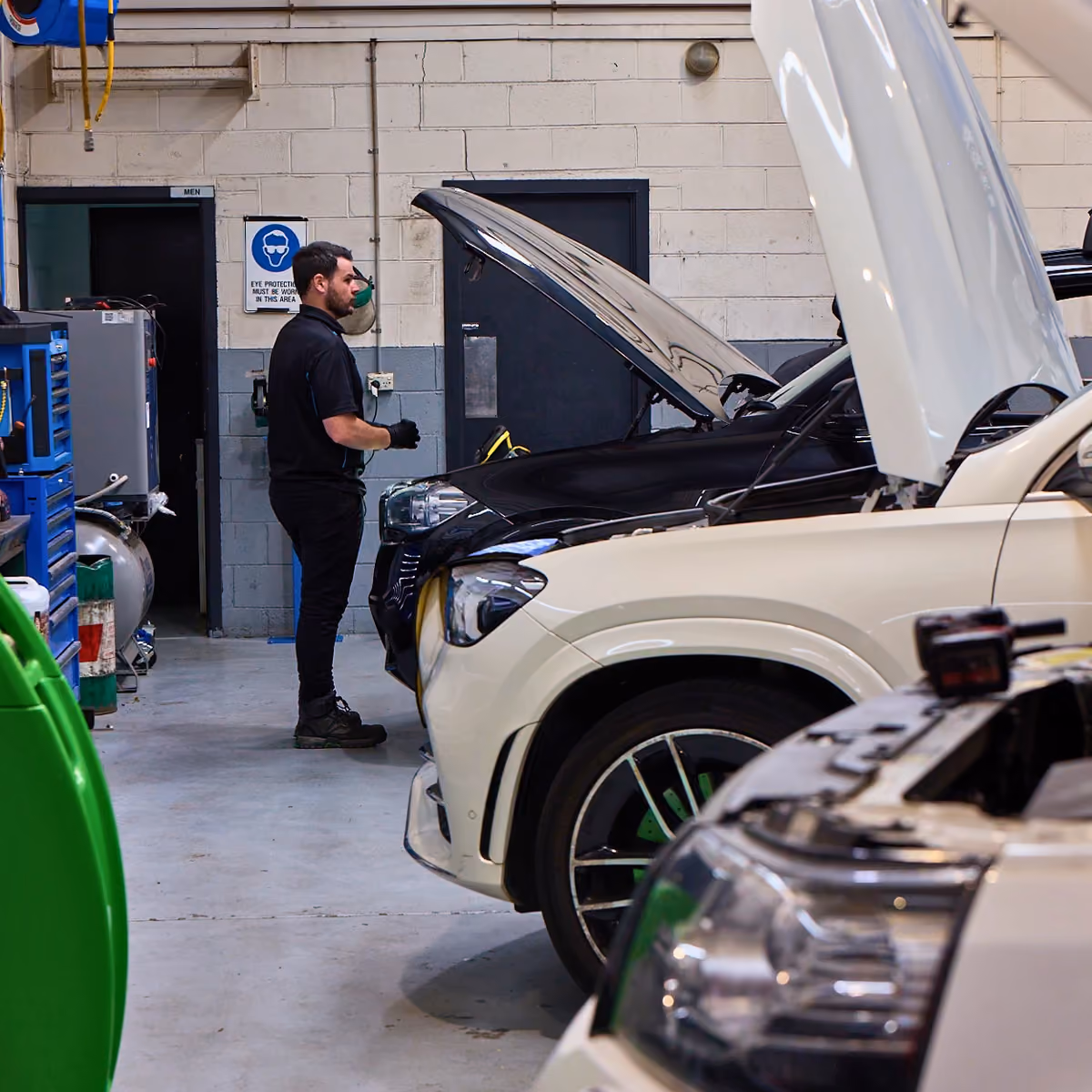 Technician inspecting vehicles with open bonnets inside workshop.