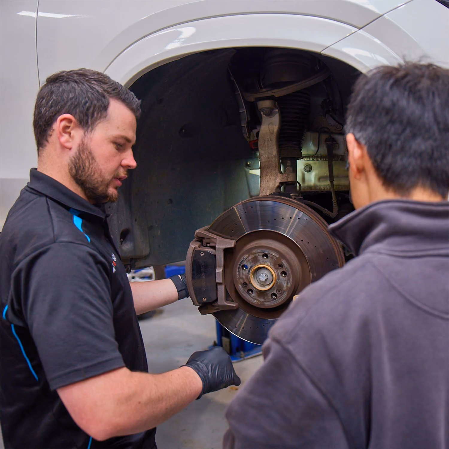 Automotive technician explaining a brake inspection on a white Lexus SUV with the front wheel removed.