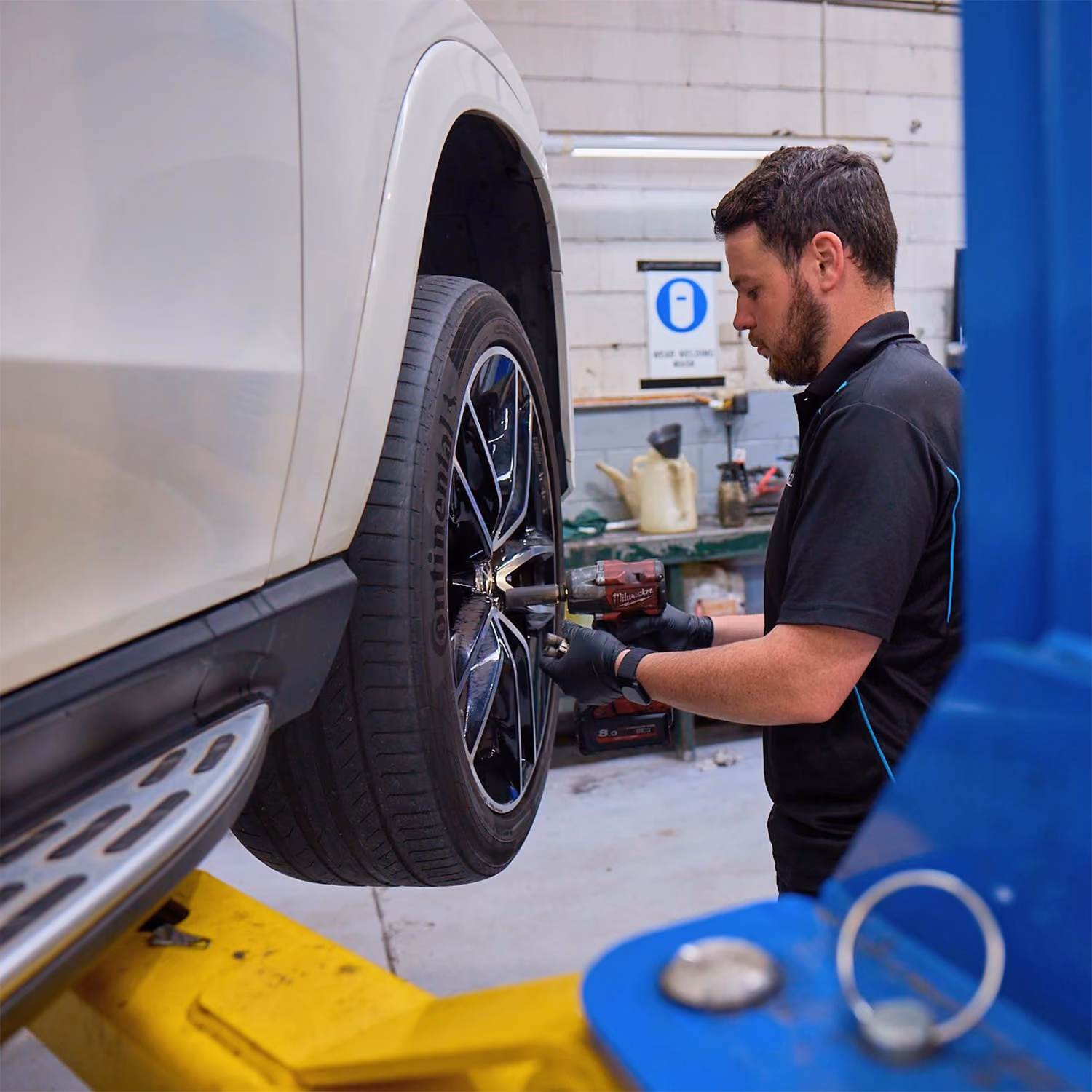 Technician performing a wheel service on a white SUV using an impact wrench while the vehicle is raised on a workshop hoist.