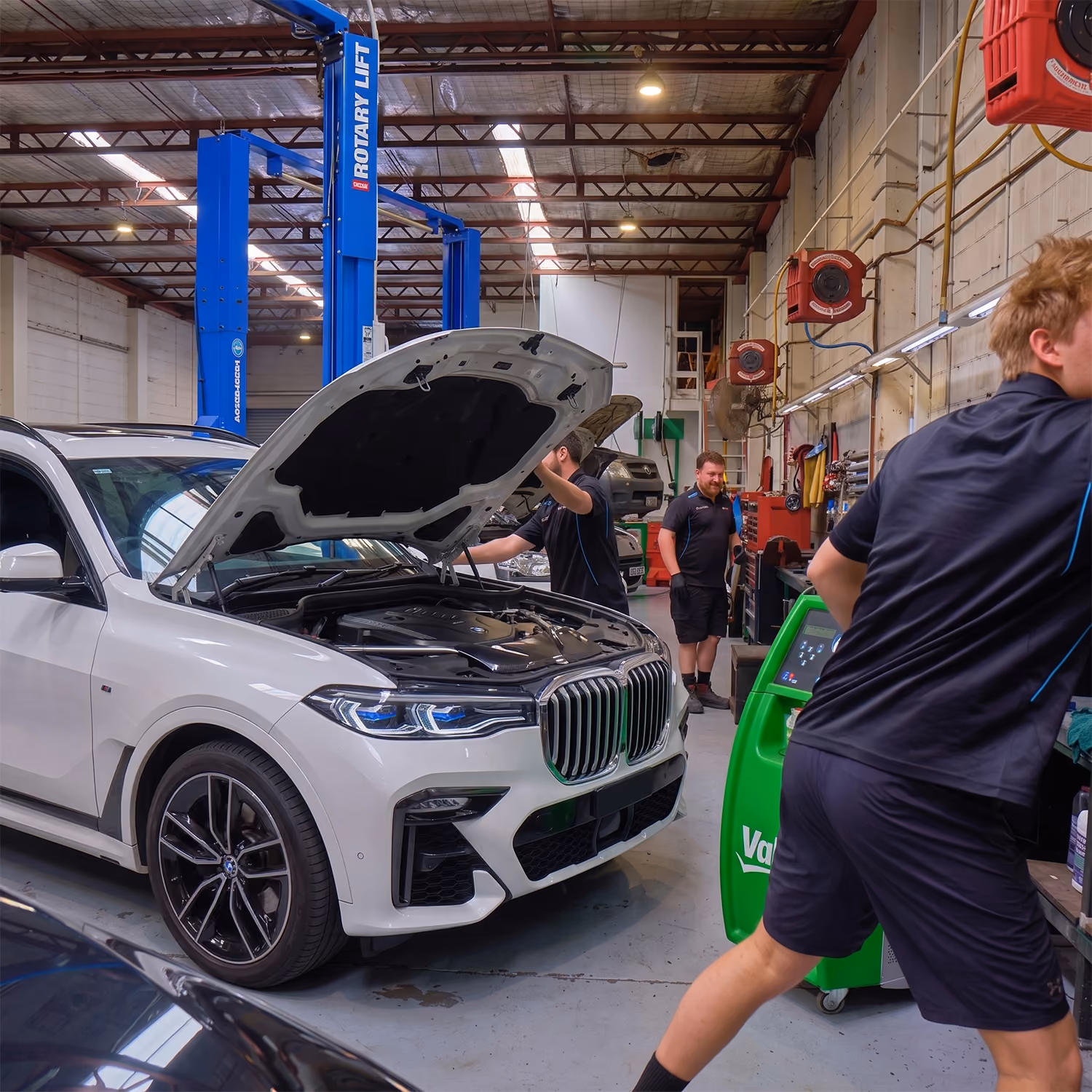 A mechanic near a green machine, next to a white BMW SUV on a lift with its hood up.