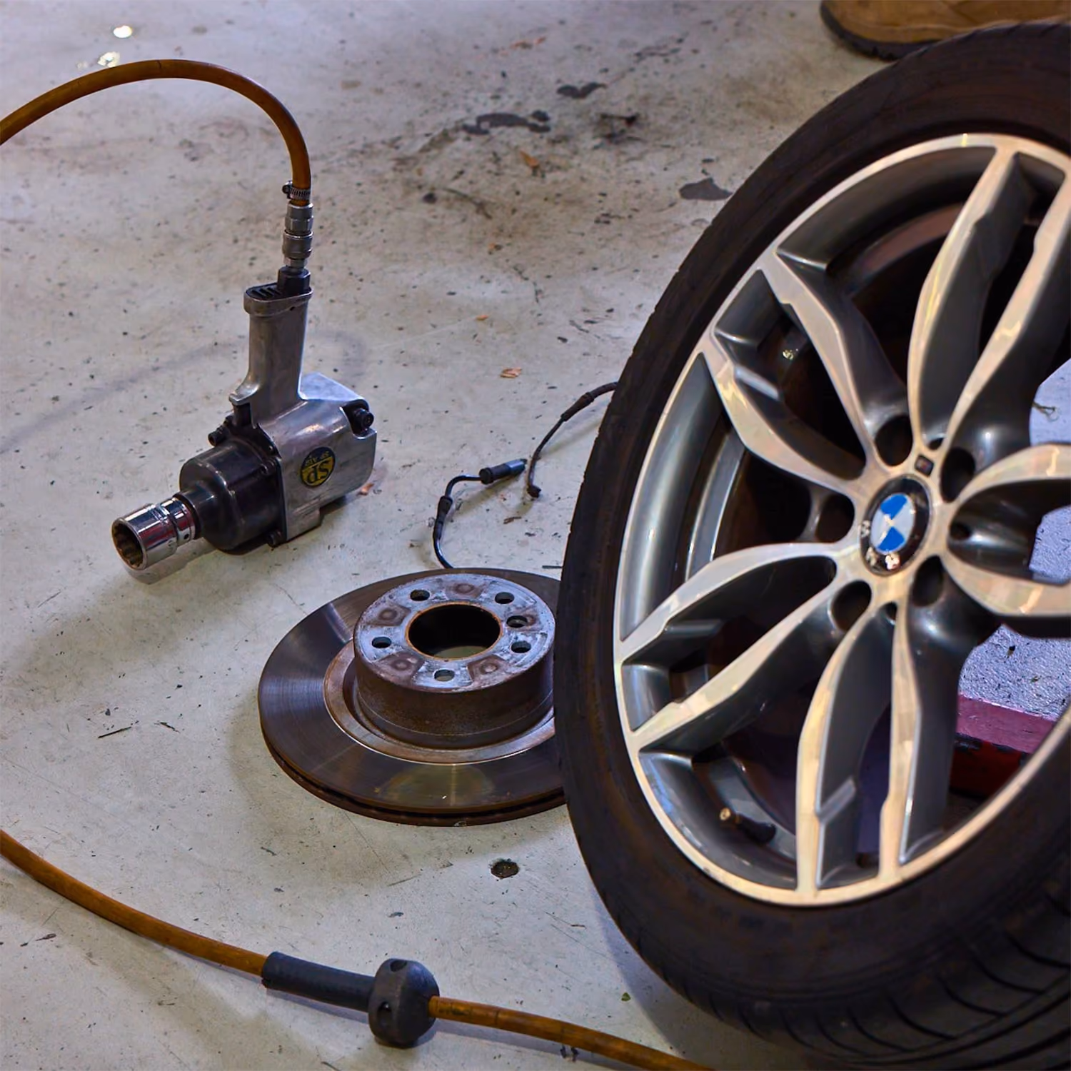 BMW alloy wheel removed during a brake service, with a brake disc, pneumatic impact wrench and workshop tools on the garage floor.