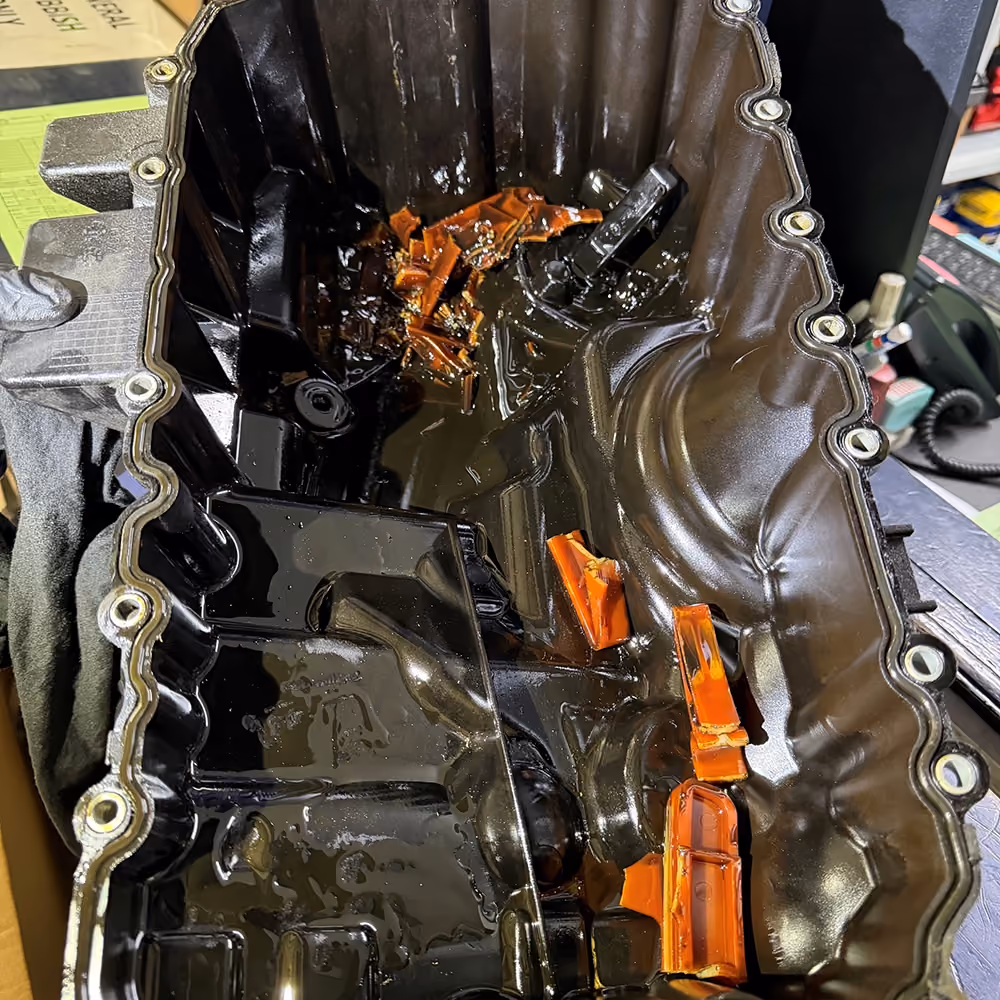 Engine oil pan containing broken orange timing chain guide fragments in used oil.
