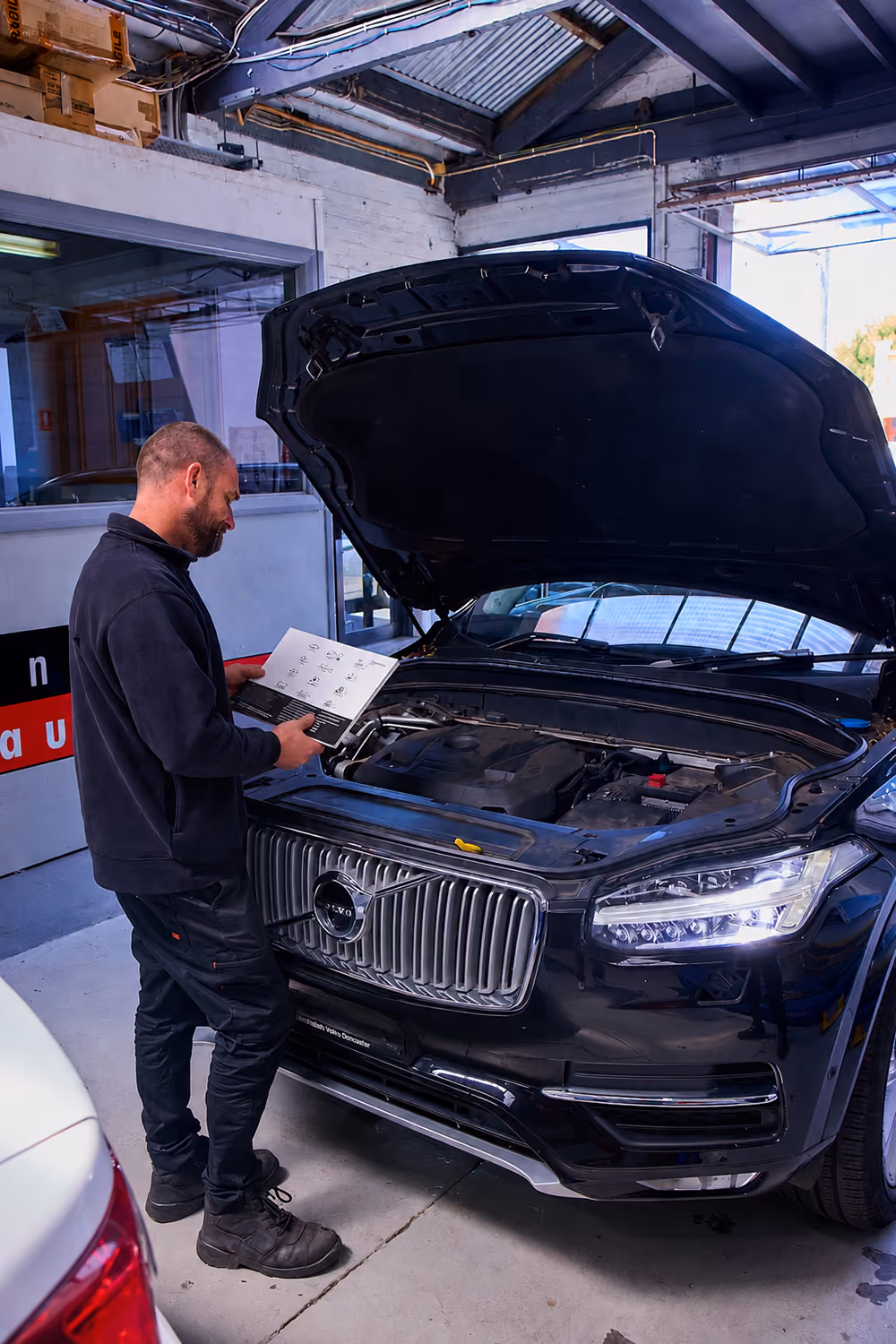 Technician reviewing a vehicle service handbook while inspecting the engine bay of a Volvo SUV with the bonnet open in an automotive workshop.