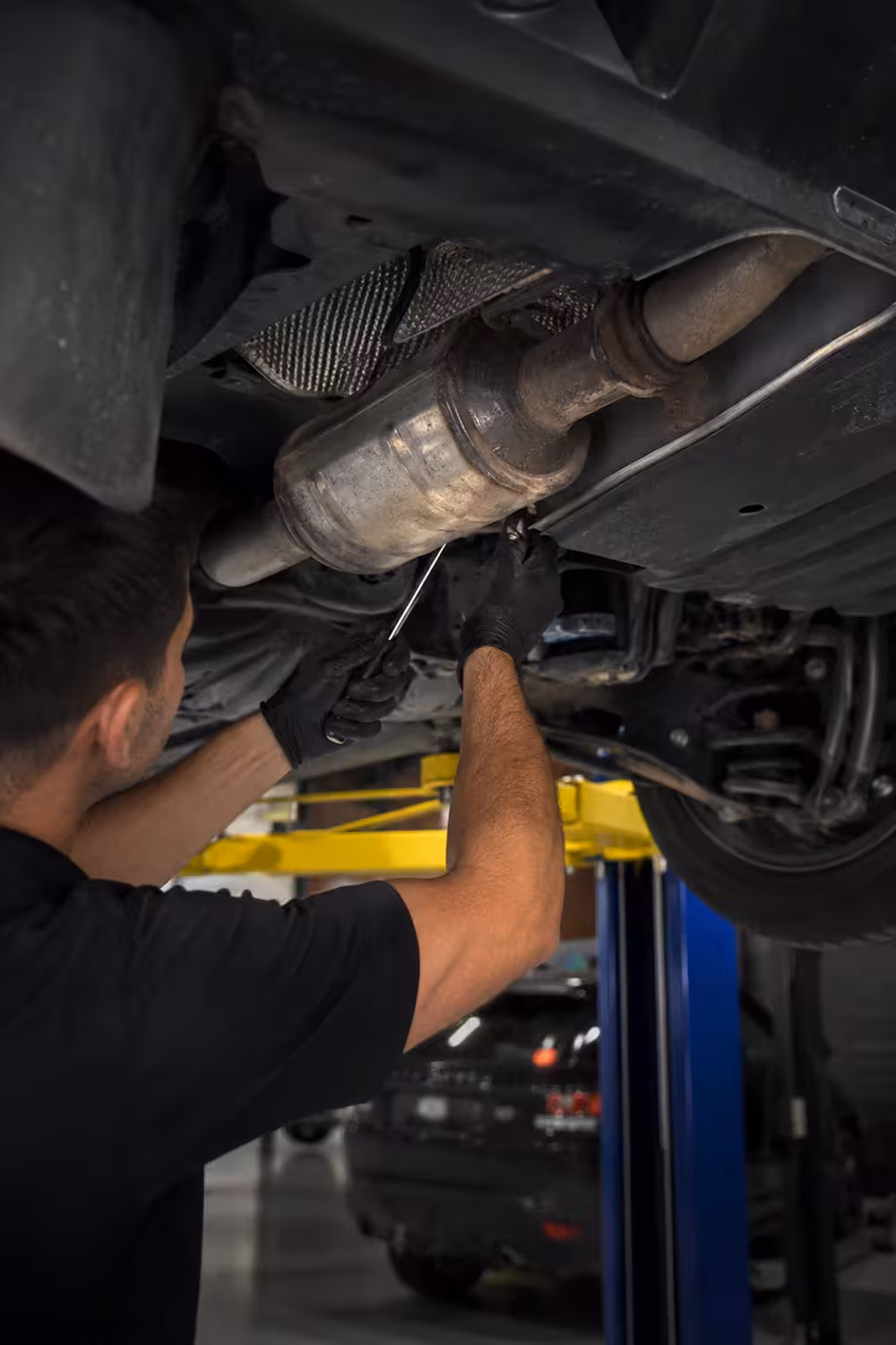 Technician inspecting and repairing a vehicle's exhaust system while working underneath a car on a hoist
