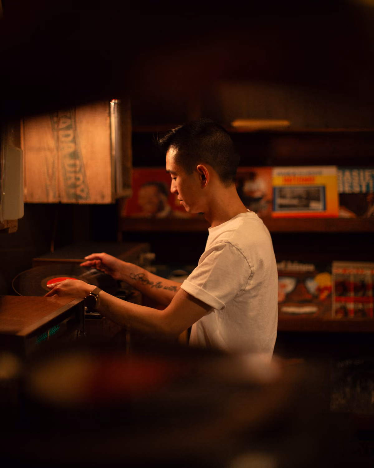 A candid photo of the candidate, Austin Seng, inside the music section of his vintage shop. He is standing in front of a vintage turntable and placing a record on it to play.