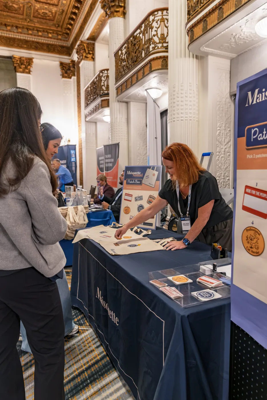 A Maisonette host laying out patches on totes for two guests at a conference booth