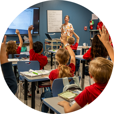 Students raising hands in a classroom while teacher stands at whiteboard