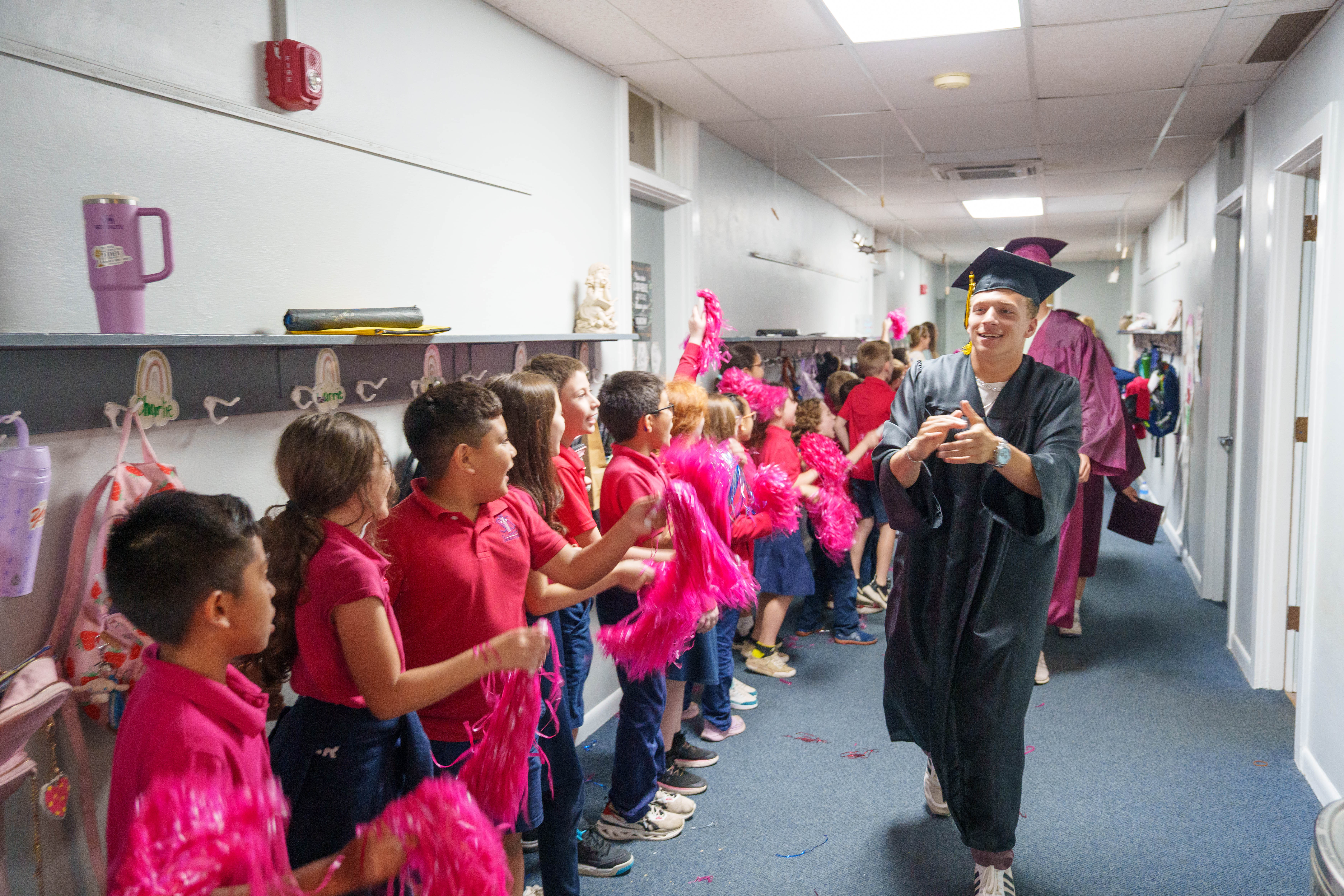 Graduate in cap and gown celebrated by elementary students waving pink pom-poms