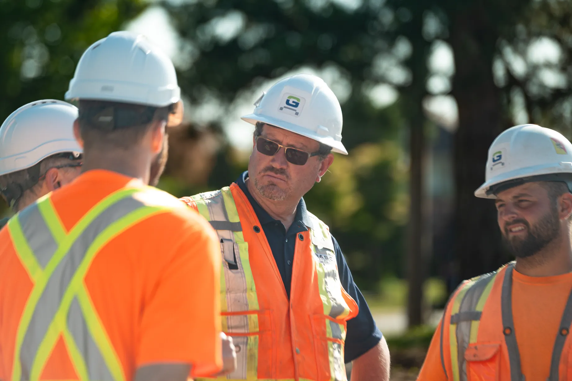 Construction workers wearing white helmets and orange safety vests engaged in discussion outdoors.