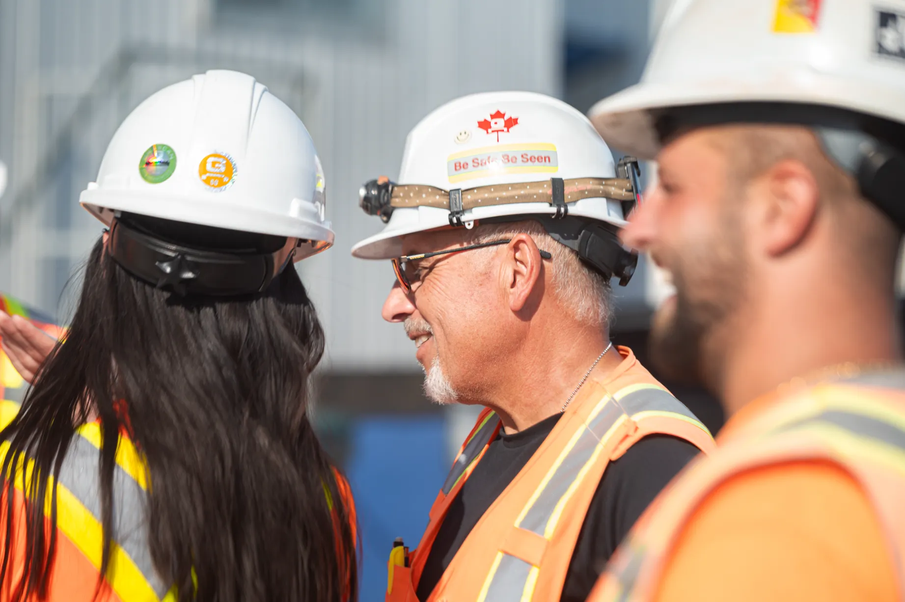 Three construction workers wearing white hard hats and orange safety vests engaged in conversation outdoors.