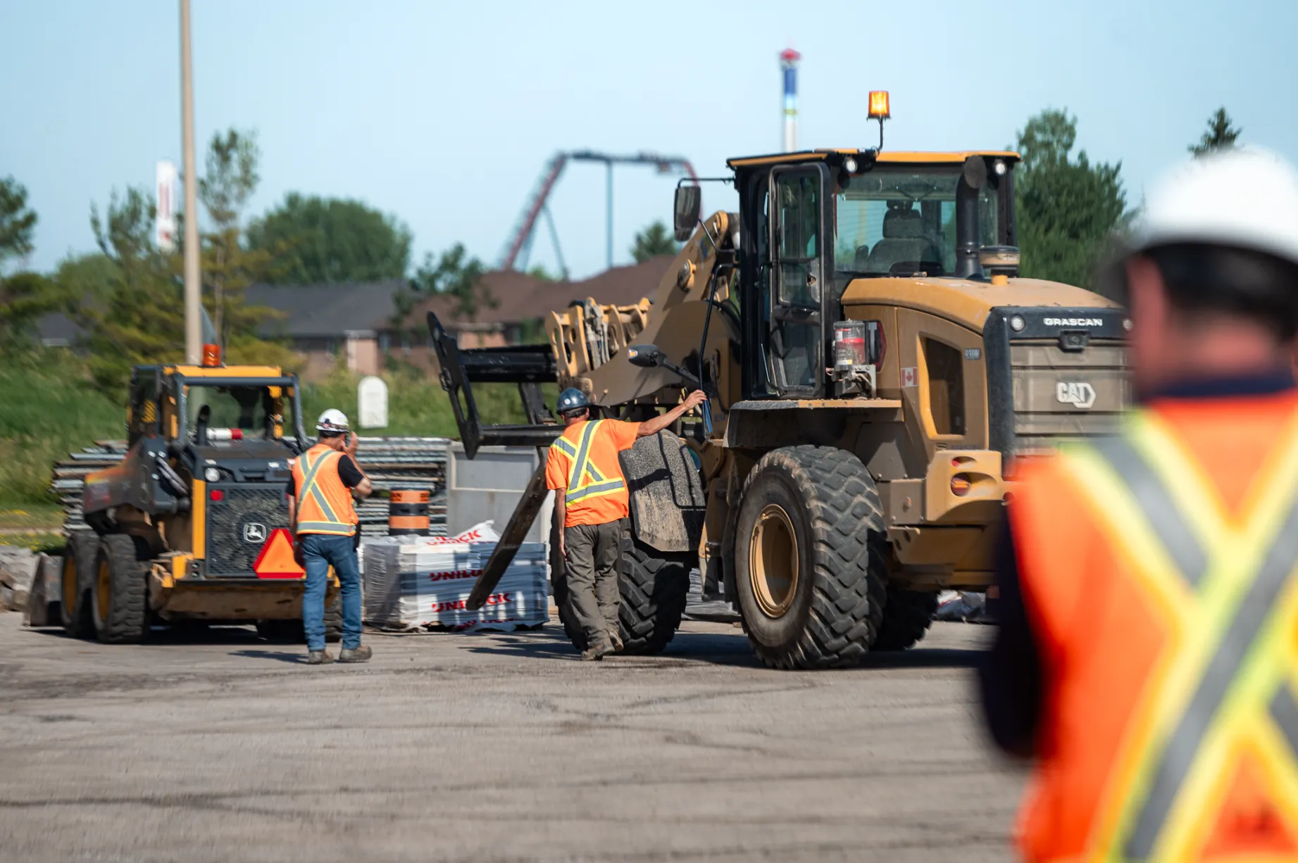 Construction workers in orange safety vests and helmets operating heavy machinery on a paved construction site.