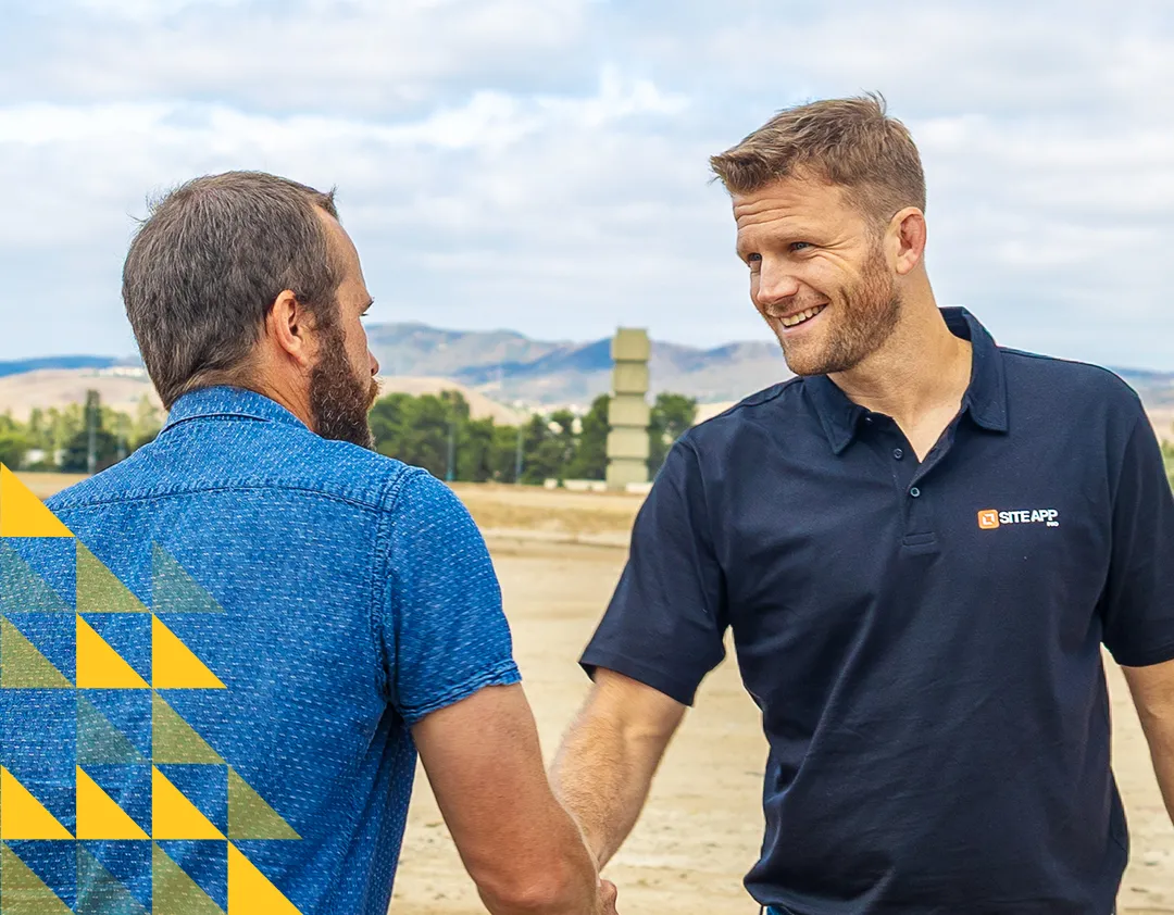 Two men shaking hands outdoors, one wearing a black polo with Site App logo and the other in a blue shirt, with mountains and trees in the background.
