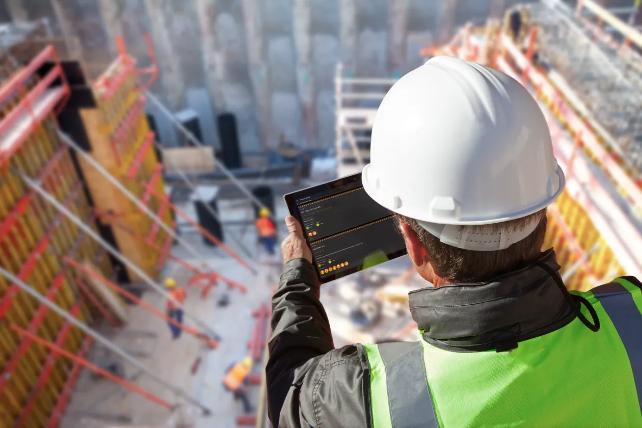 Construction worker wearing a white hard hat and green safety vest using a tablet device at a building site.