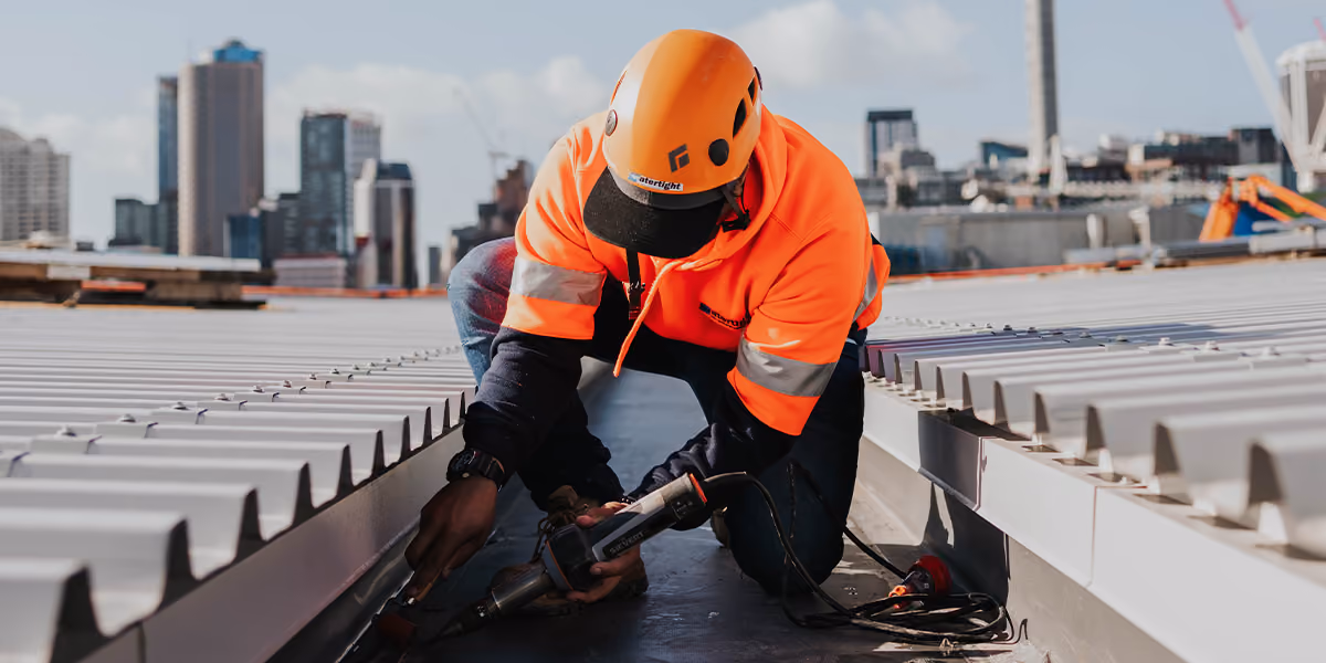 Roofer in hi-vis kneeling on a metal roof using a power tool, city skyline behind