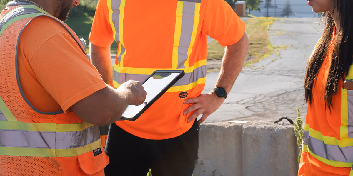 Construction workers in orange hi-vis vests gathered around a tablet at a roadside worksite