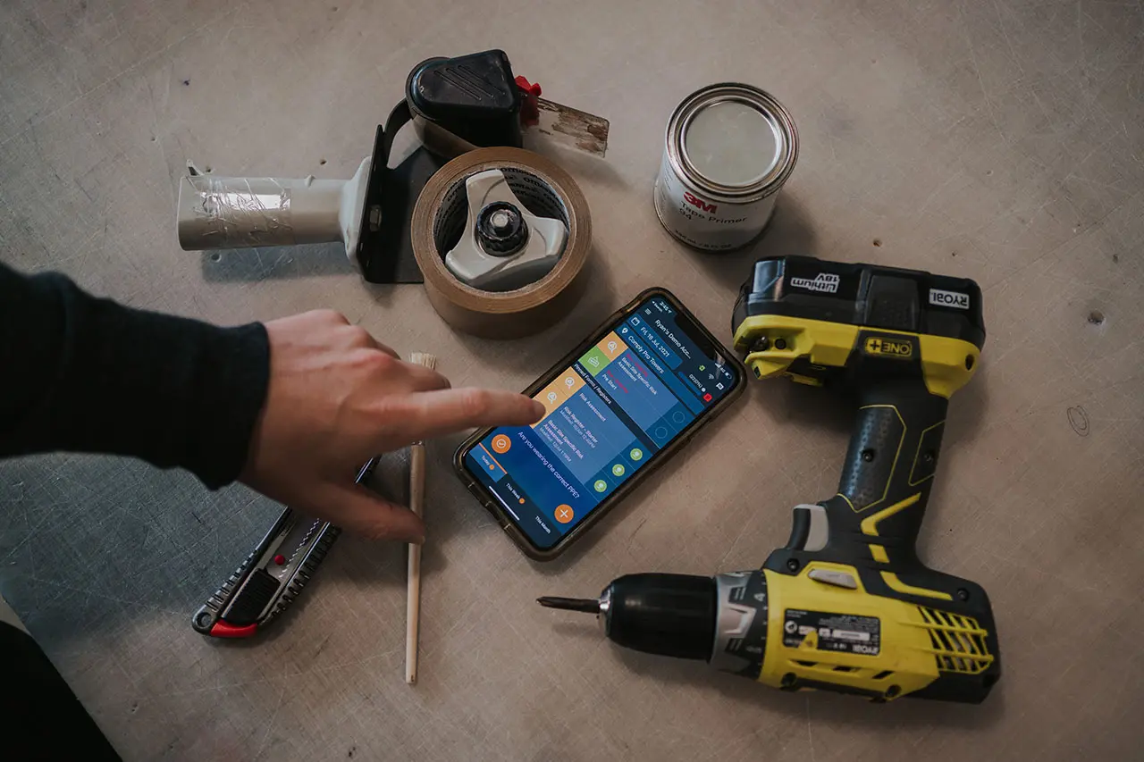 Hand reaching for a phone showing a safety app among a drill, tape and tools on a concrete floor