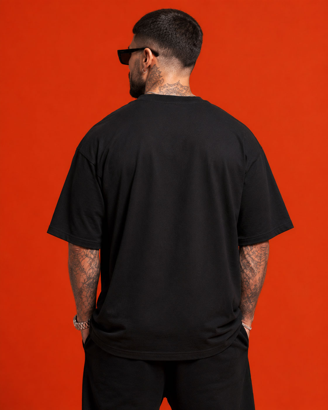 Man with tattoos on neck and arms wearing black t-shirt and sunglasses against red background, back view.