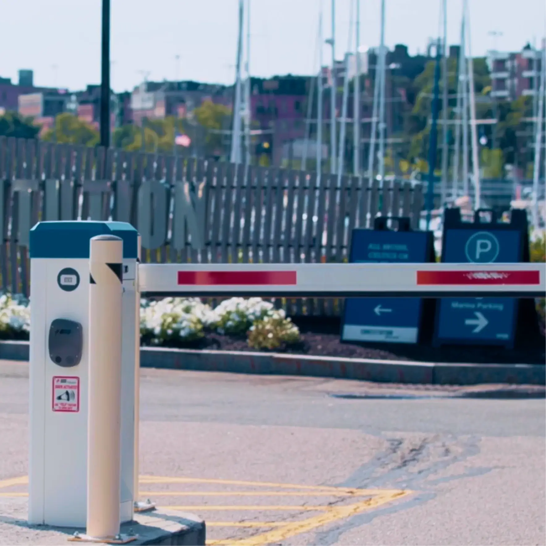 Automatic parking gate arm lowered at a parking lot entrance with signs and boats in background.