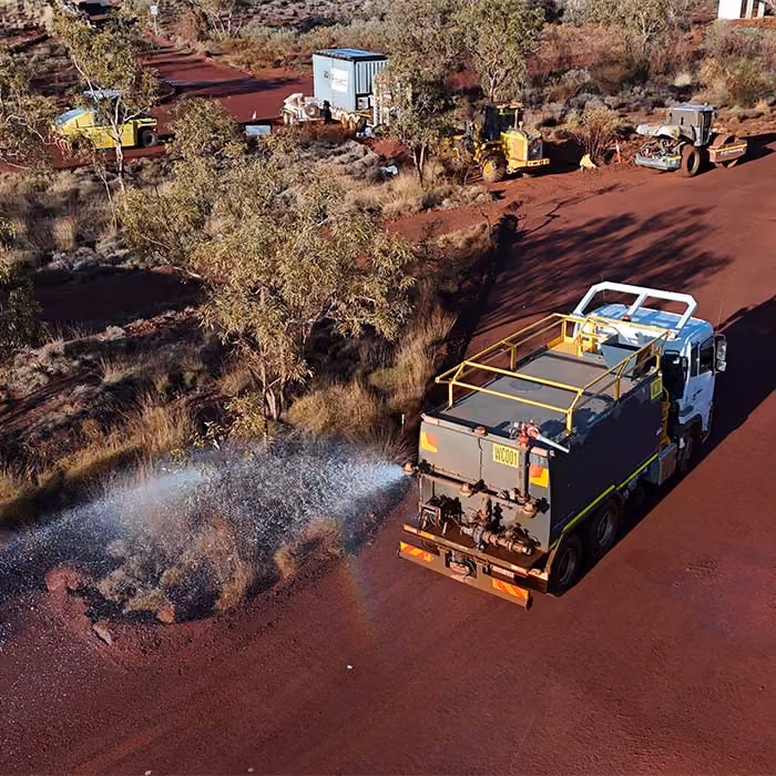 Two dump trucks and a road roller paving asphalt on a remote highway near dry hills and railway tracks.