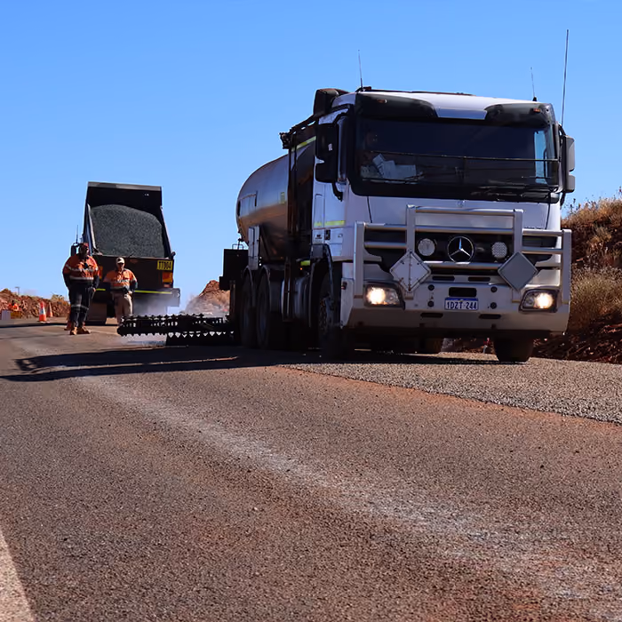 Two dump trucks and a road roller paving asphalt on a remote highway near dry hills and railway tracks.