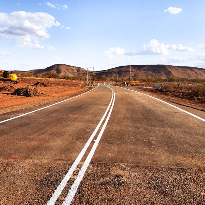 Two dump trucks and a road roller paving asphalt on a remote highway near dry hills and railway tracks.
