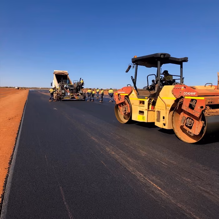 Two dump trucks and a road roller paving asphalt on a remote highway near dry hills and railway tracks.