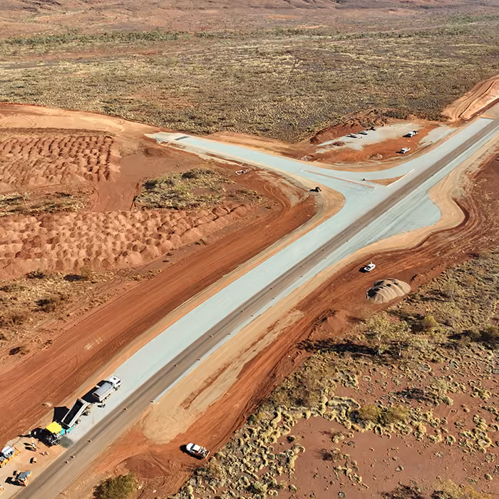 Two dump trucks and a road roller paving asphalt on a remote highway near dry hills and railway tracks.