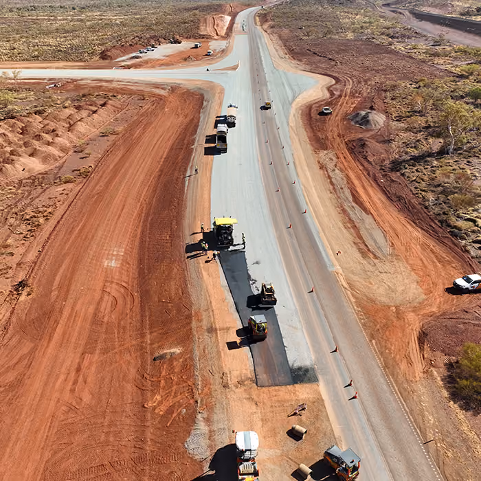 Two dump trucks and a road roller paving asphalt on a remote highway near dry hills and railway tracks.